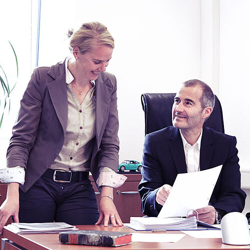 Ein sitzender Mann zeigt der stehenden Frau auf einem Papier etwas.