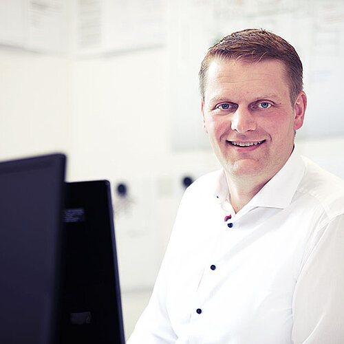 Mann im weißen Hemd sitzt am Schreibtisch und guckt seitlich in die Kamera.