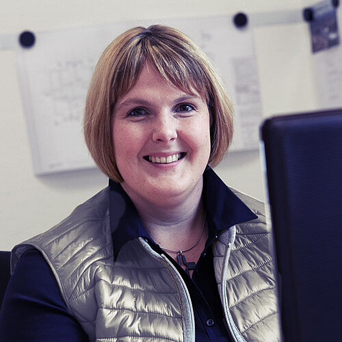 Eine Frau mit einem Bob-Haarschnitt sitzt am Arbeitsplatz und lächelt in die Kamera.