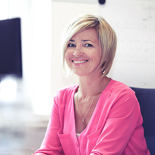 Blonde Frau mit pinker Bluse sitzt lächeln am Arbeitsplatz und guckt in die Kamera.
