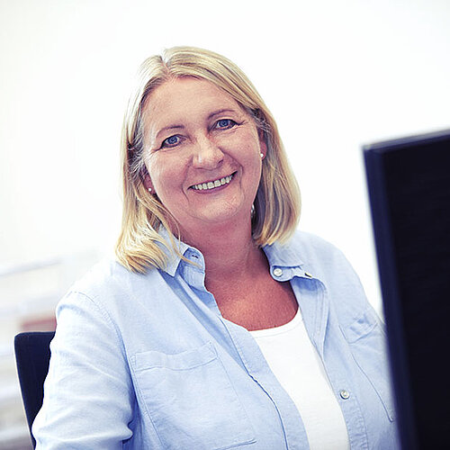 Eine blonde Frau mit hellblauer Bluse sitzt am Schreibtisch und schaut lächelnd in die Kamera.